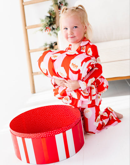 A young girl with blonde pigtails sits on a white floor next to a large red and white striped gift box, wearing a red and white Santa Claus patterned bamboo pajama set in a bright, decorated room.