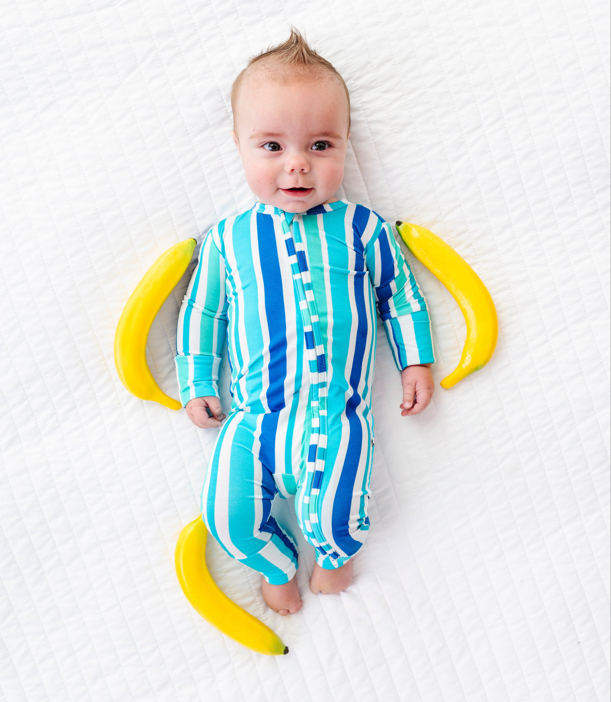 A baby lying on a white textured blanket, wearing a blue and turquoise striped one-piece jumpsuit. The baby is surrounded by two yellow bananas, one on each side.