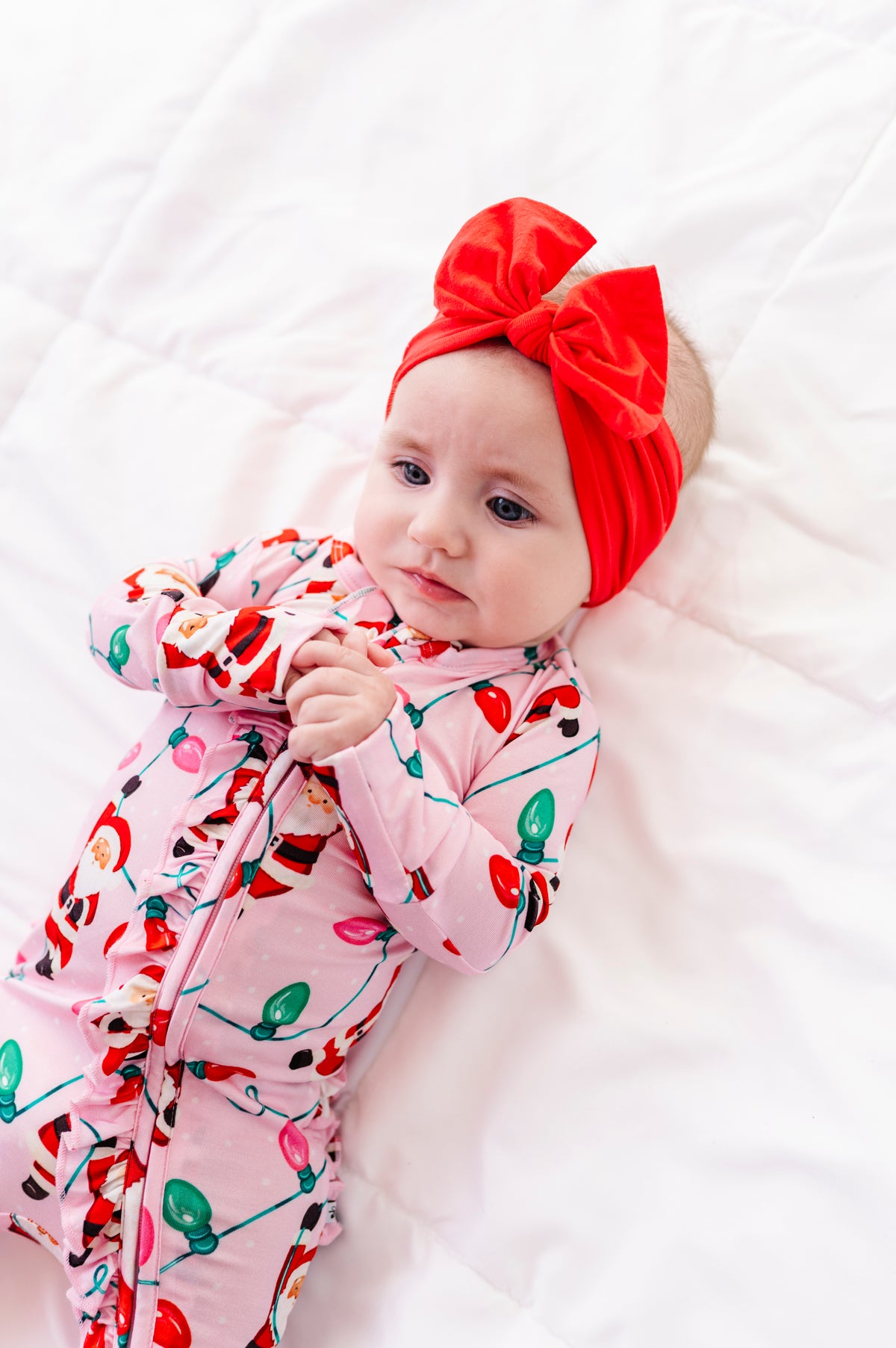 A baby girl wearing a pink ruffle bamboo convertible romper featuring  variety of Santas hanging from multicolor Christmas lights 