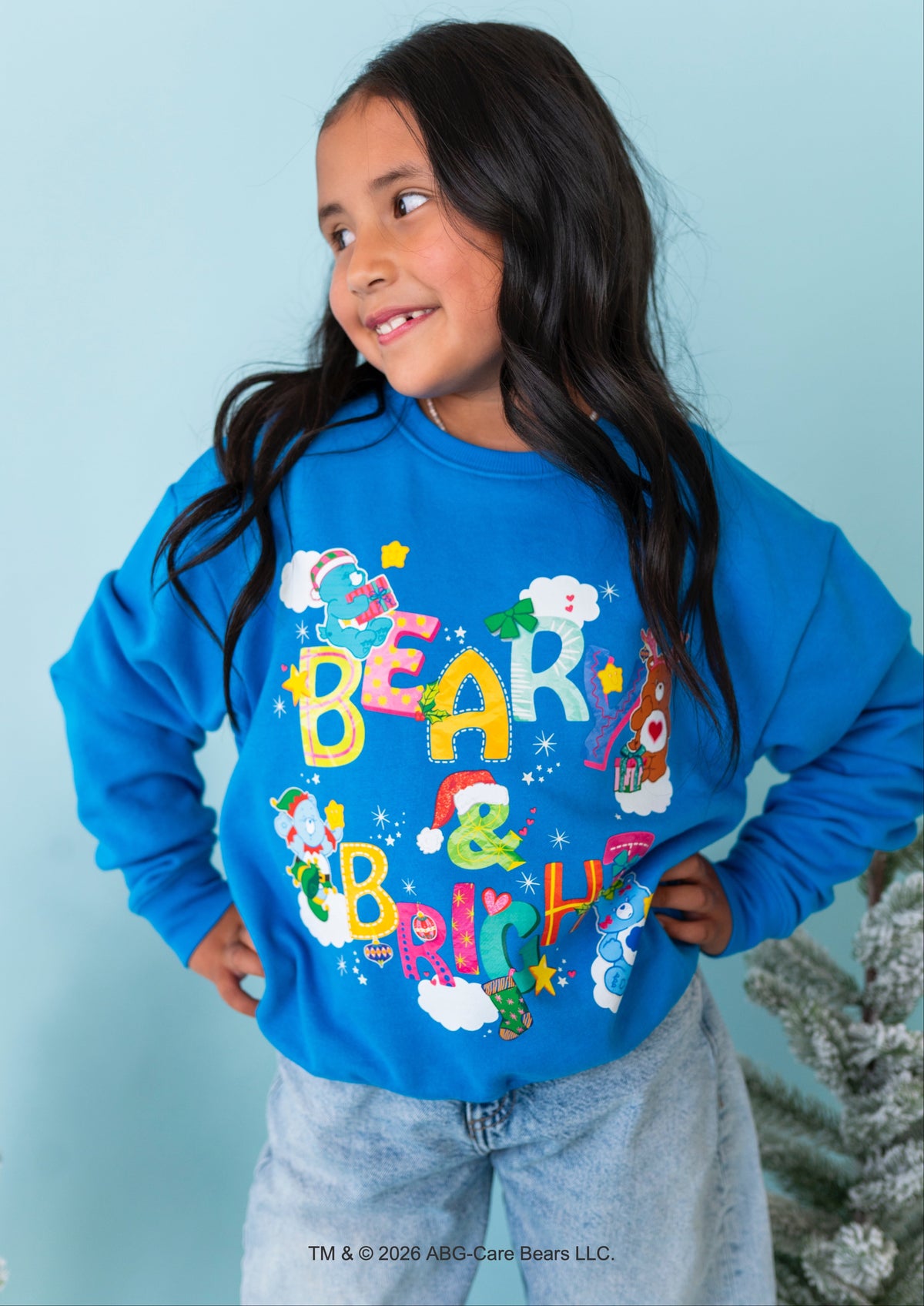 A young girl with long dark hair wearing a bright blue sweatshirt with a colorful 'Bear & Brito' Christmas graphic, paired with light wash jeans, standing against a light blue wall.