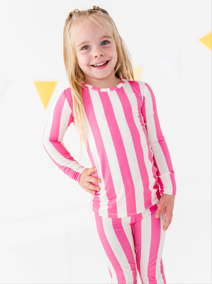 A young girl with blonde hair in buns is smiling and posing in a pink and white vertically striped long-sleeve rash guard and matching striped swim leggings against a white background with subtle yellow triangle decorations.