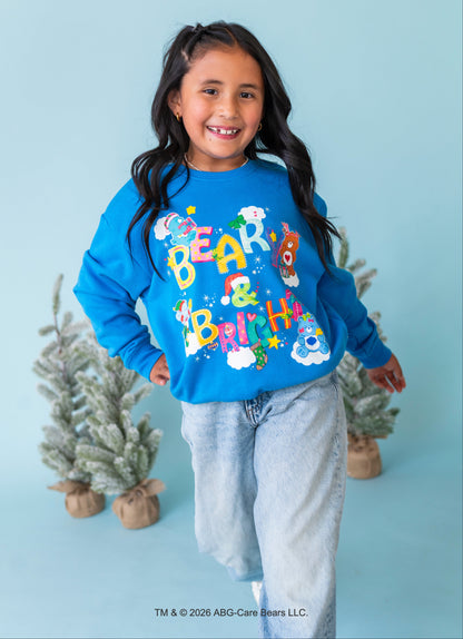 A young girl smiling and posing in a bright blue crewneck sweatshirt with a colorful 'Bear & Bunny' graphic, paired with light-wash jeans, set against a light blue background with small decorative trees.