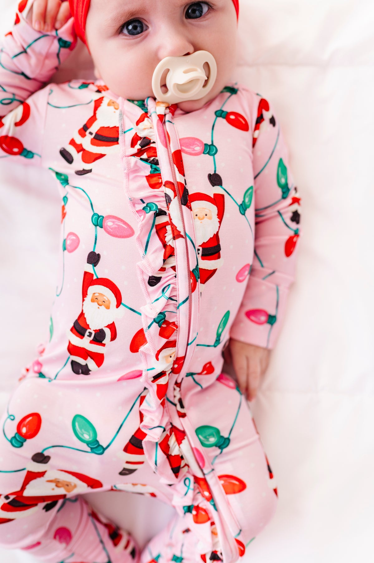 A close-up detail shot of a baby girl wearing a pink ruffle bamboo convertible romper featuring  variety of Santas hanging from multicolor Christmas lights 