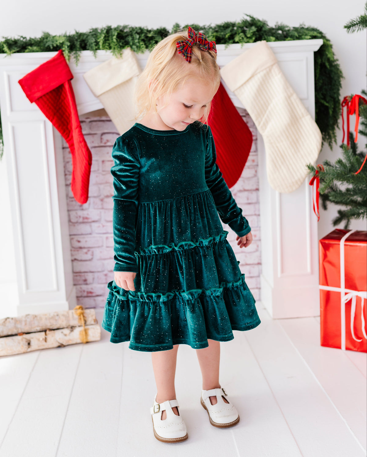 A toddler girl wearing a dark green velvet long-sleeve tiered dress and white shoes, standing in front of a decorated Christmas fireplace with stockings, greenery, and wrapped gifts.