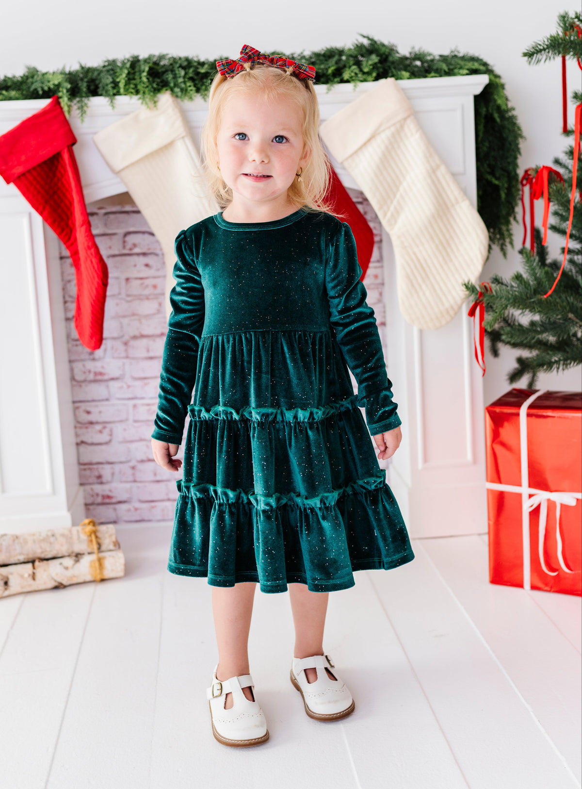 A young toddler girl stands in front of a white fireplace mantel decorated with Christmas stockings, greenery, and a tree, wearing a long-sleeve dark green velvet tiered dress and white shoes.