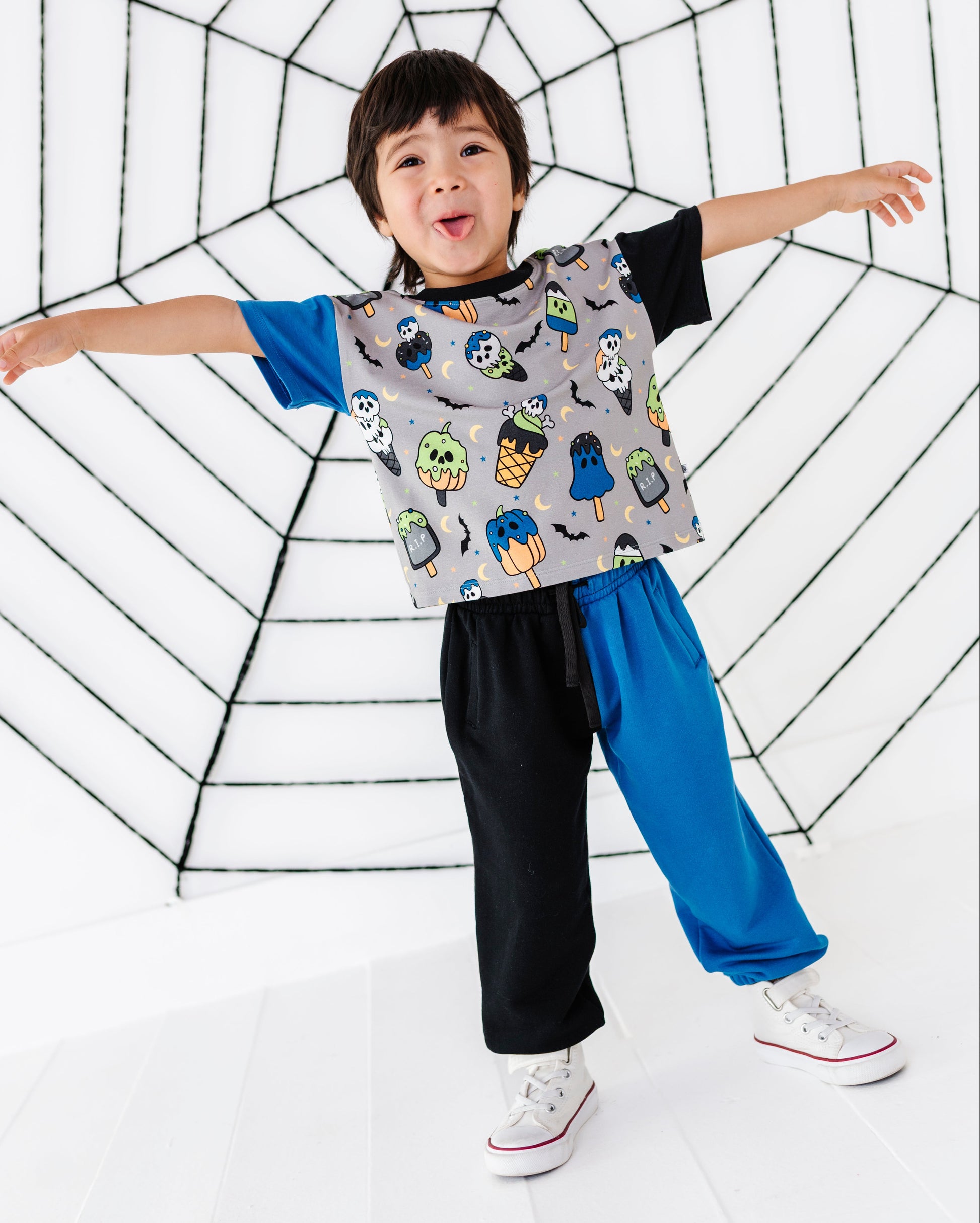 A young boy wearing a gray short-sleeve t-shirt with a colorful owl pattern, paired with black and blue sweatpants and white sneakers, posing with outstretched arms in front of a large black spiderweb structure on a white background.