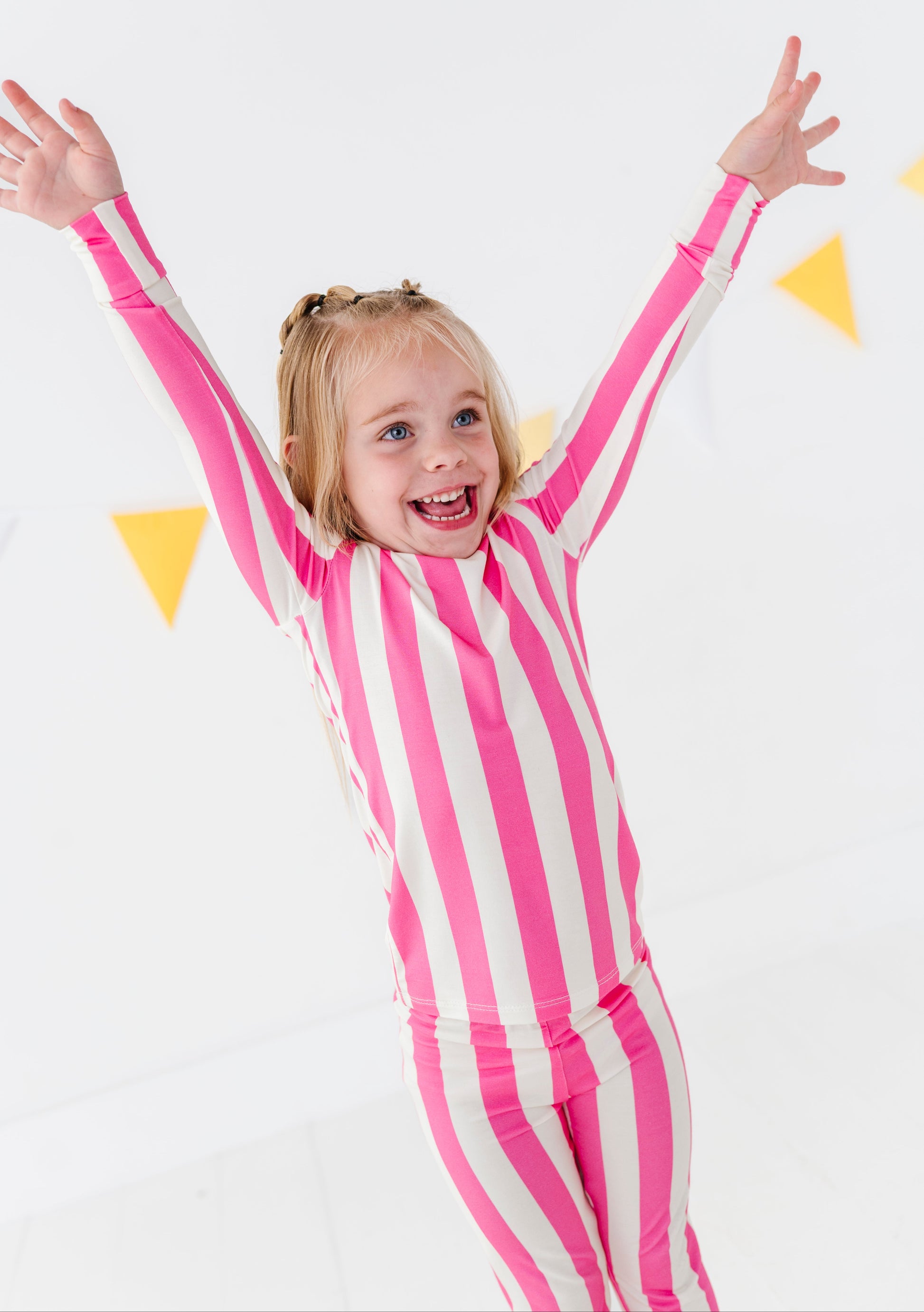 A young girl with blonde hair in a ponytail is wearing a pink and white vertically striped long-sleeve bodysuit, smiling and raising her arms against a white background with yellow triangular decorations.