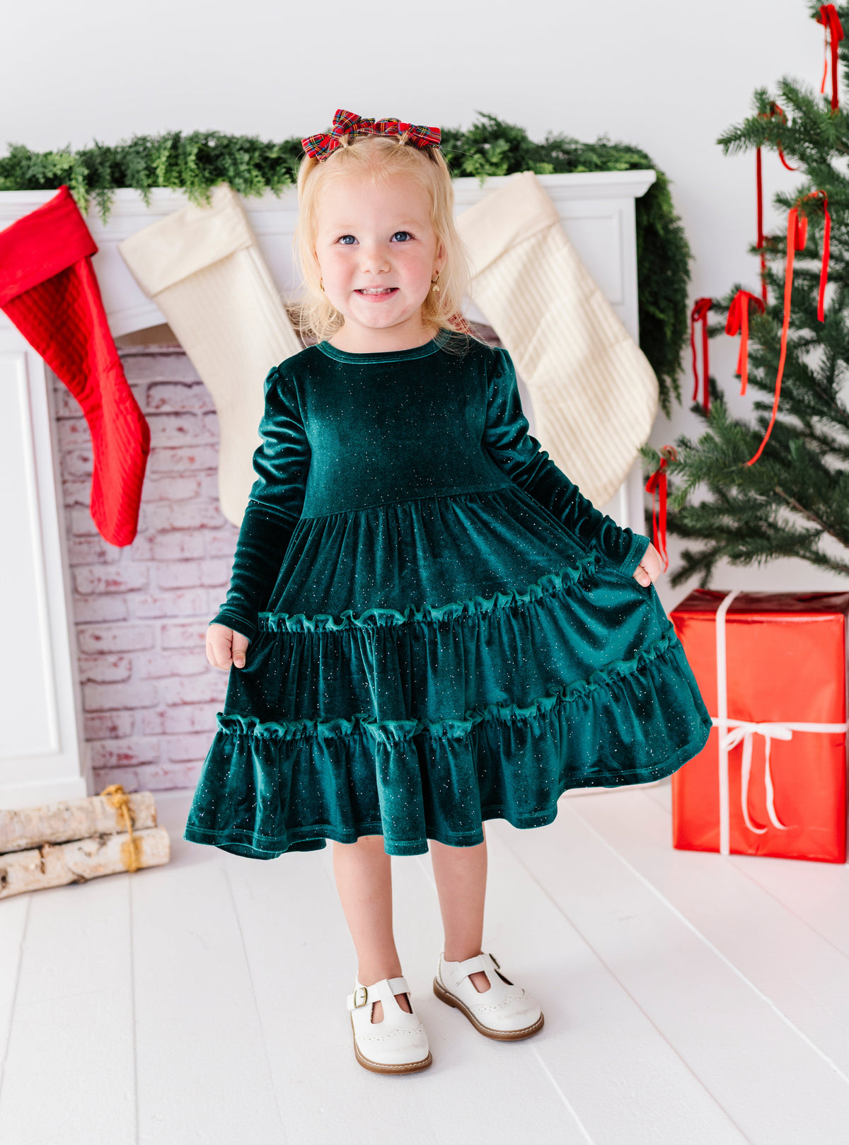 A young girl stands in a festive holiday setting wearing a dark green velvet long-sleeve tiered dress with white shoes. The background features a decorated fireplace with stockings, a Christmas tree, and wrapped gifts.