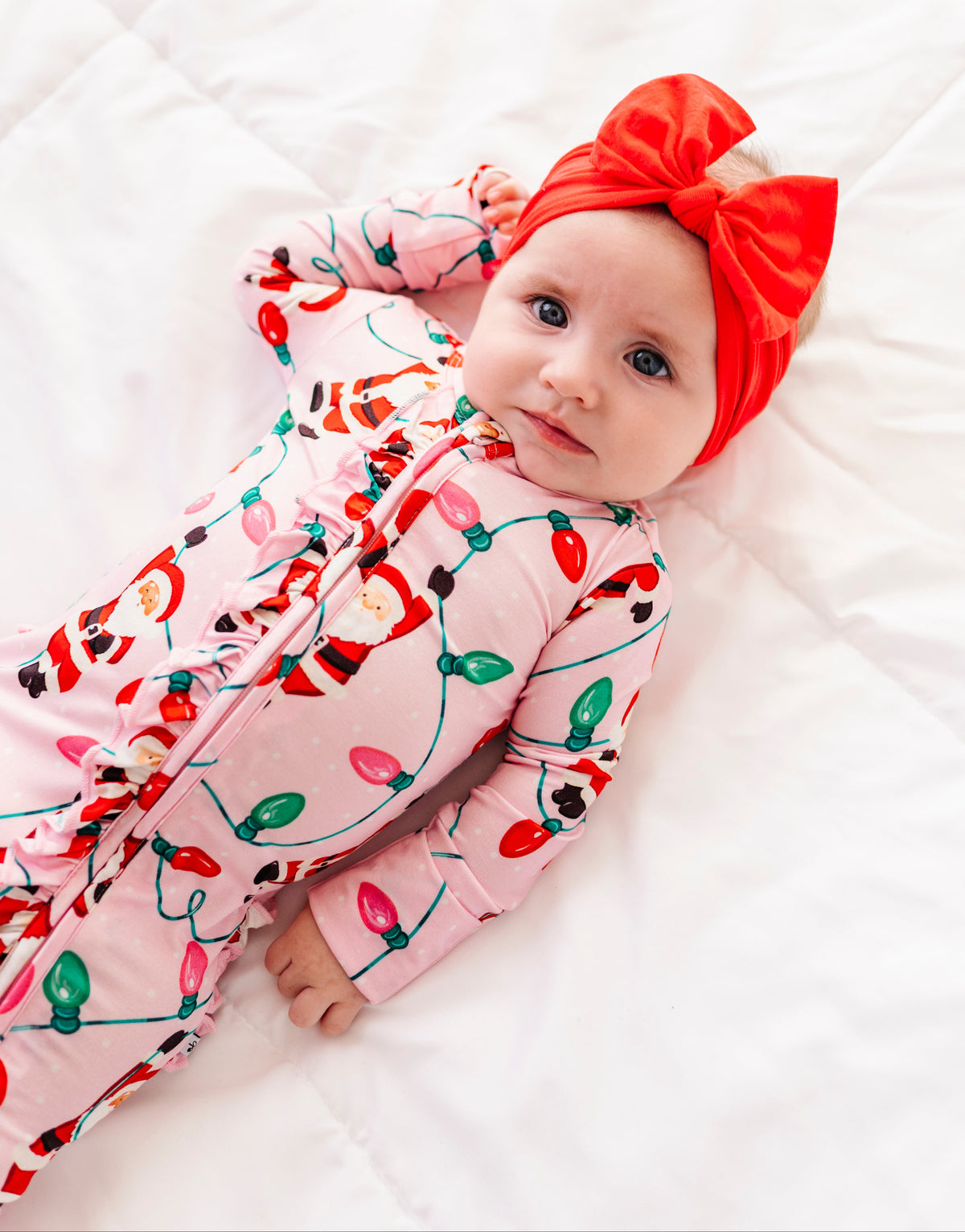 A baby girl wearing a pink ruffle bamboo convertible romper featuring  variety of Santas hanging from multicolor Christmas lights 