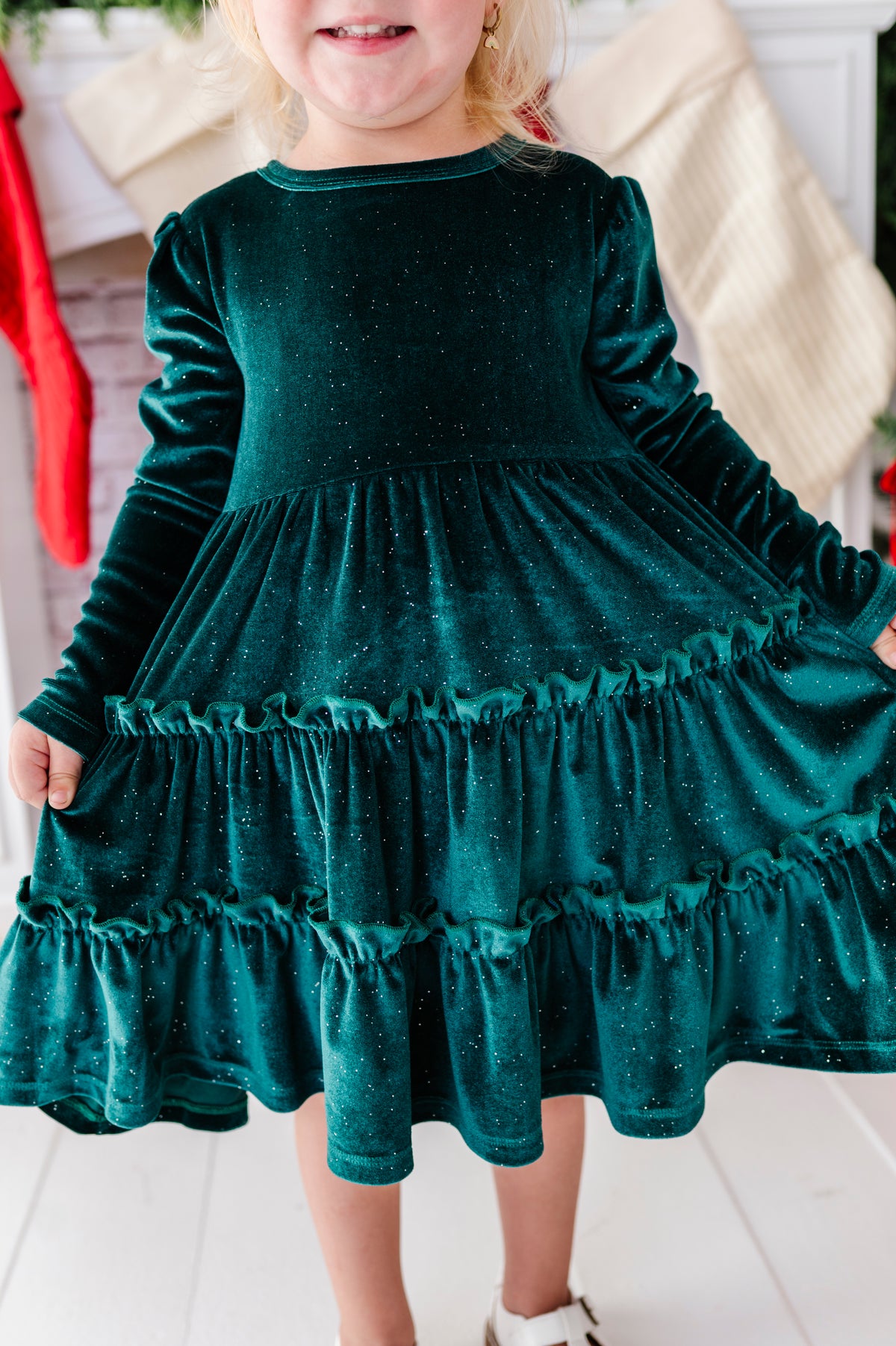 A young child wearing a sparkling dark green velvet long-sleeve dress with a tiered ruffle skirt, standing in a festive indoor setting.