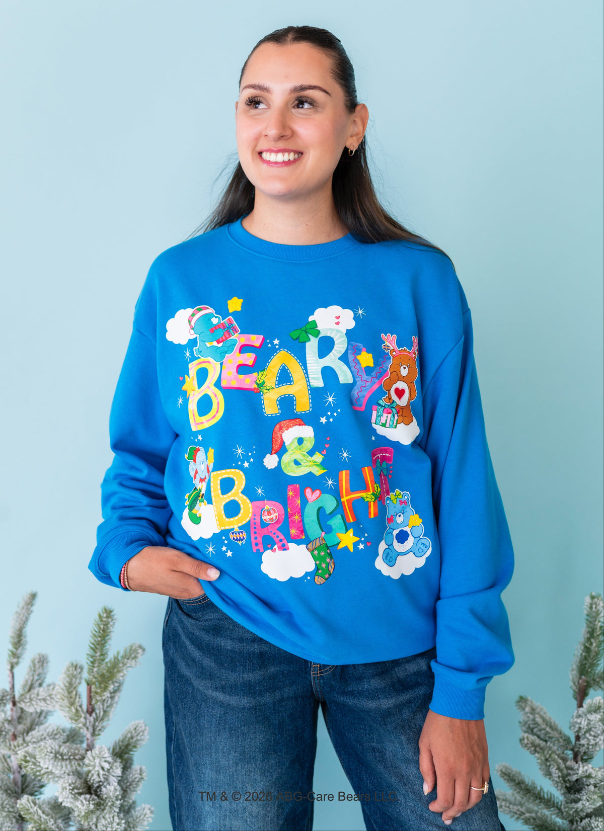 A woman wearing a bright blue crewneck sweatshirt with a colorful 'Bear & Bright' graphic featuring cartoon bears, clouds, and stars, paired with dark blue jeans, set against a light blue background with decorative pine branches.