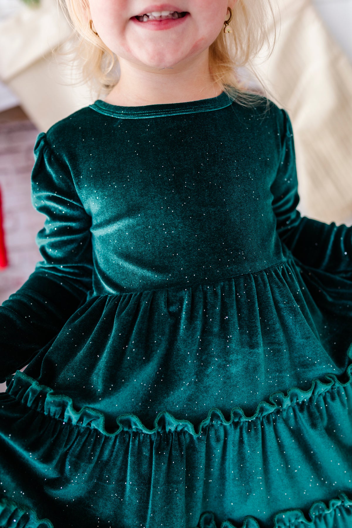 A close-up of a young child wearing a sparkling dark teal green velvet long-sleeve dress with tiered ruffles.