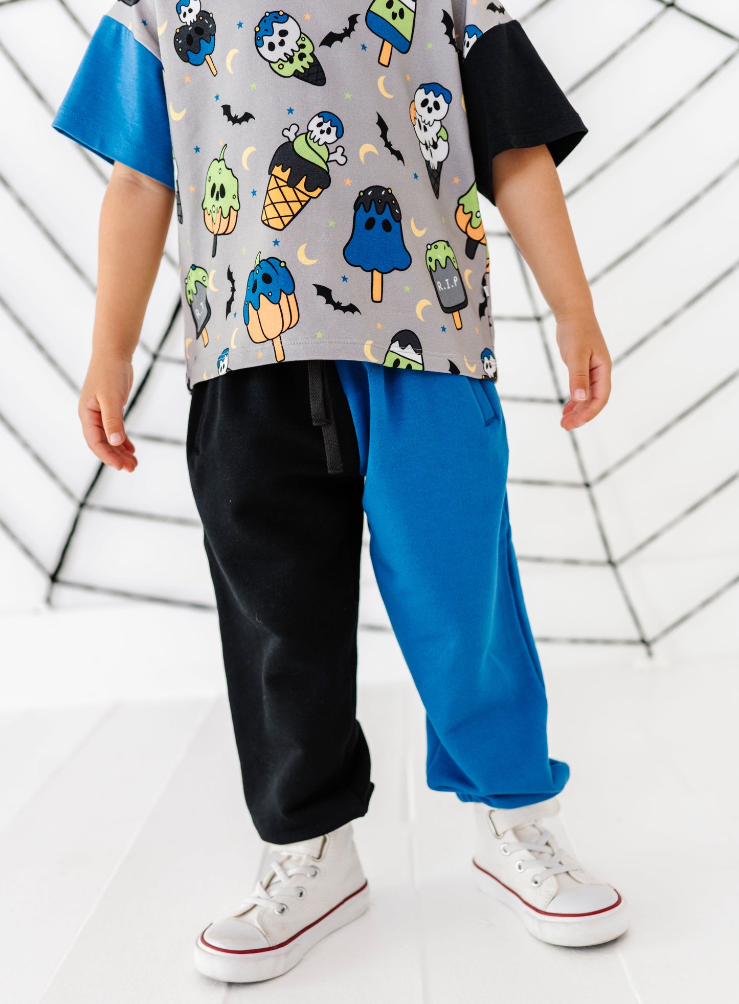 A child wearing a short-sleeve t-shirt with a colorful ice cream cone and ghost pattern on a gray base, paired with split black and bright blue pants and white sneakers, standing in front of a white web-like backdrop.