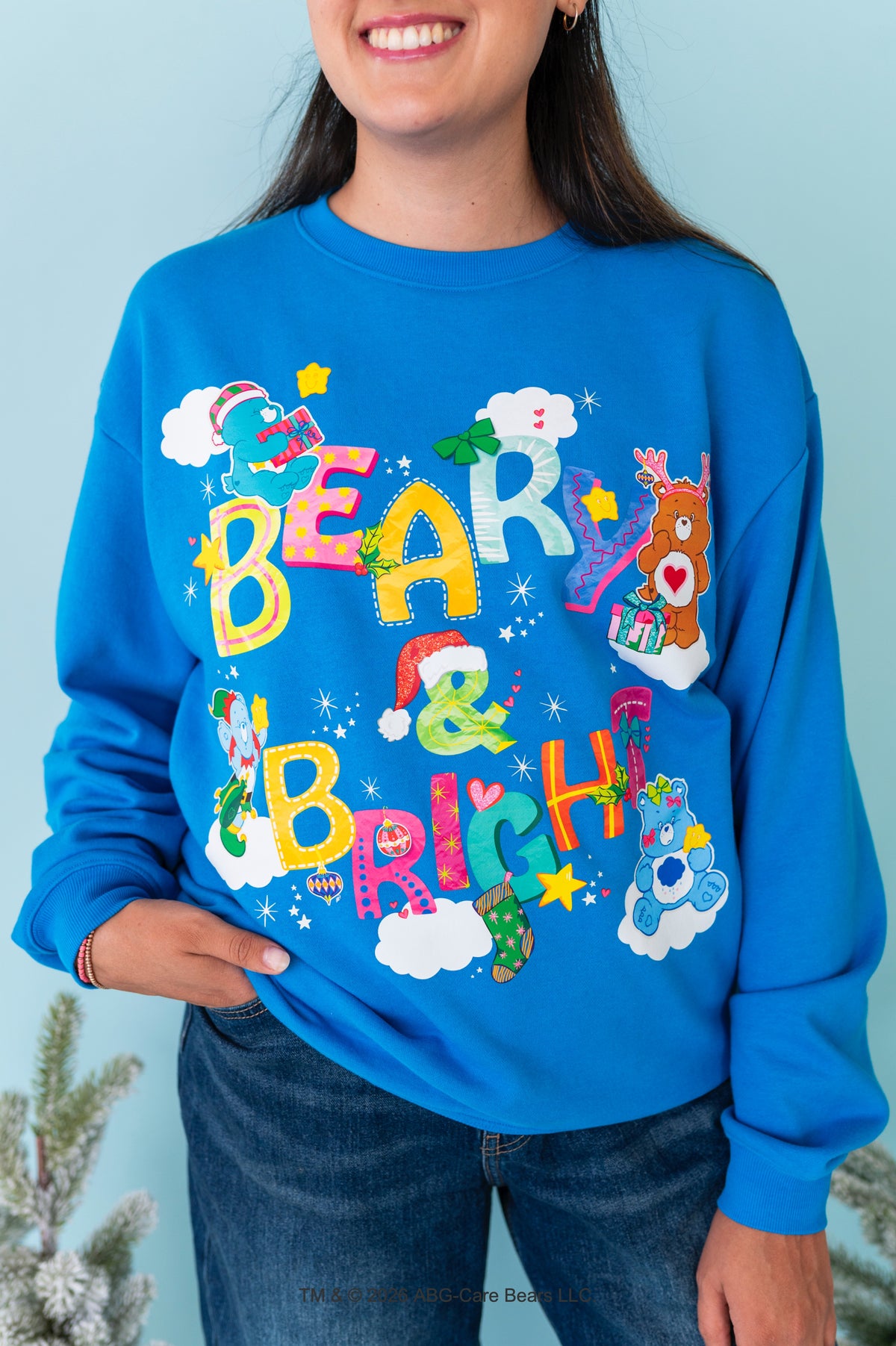 A woman wearing a bright blue crewneck sweatshirt with a colorful 'Bear & Brisket' Christmas graphic featuring cartoon bears, Santa hats, and stars, paired with dark blue jeans against a light blue background.