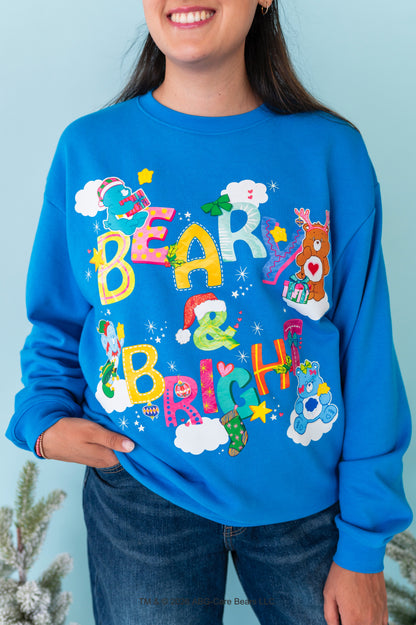 A woman wearing a bright blue crewneck sweatshirt with a colorful 'Bear & Brisket' Christmas graphic featuring cartoon bears, Santa hats, and stars, paired with dark blue jeans against a light blue background.