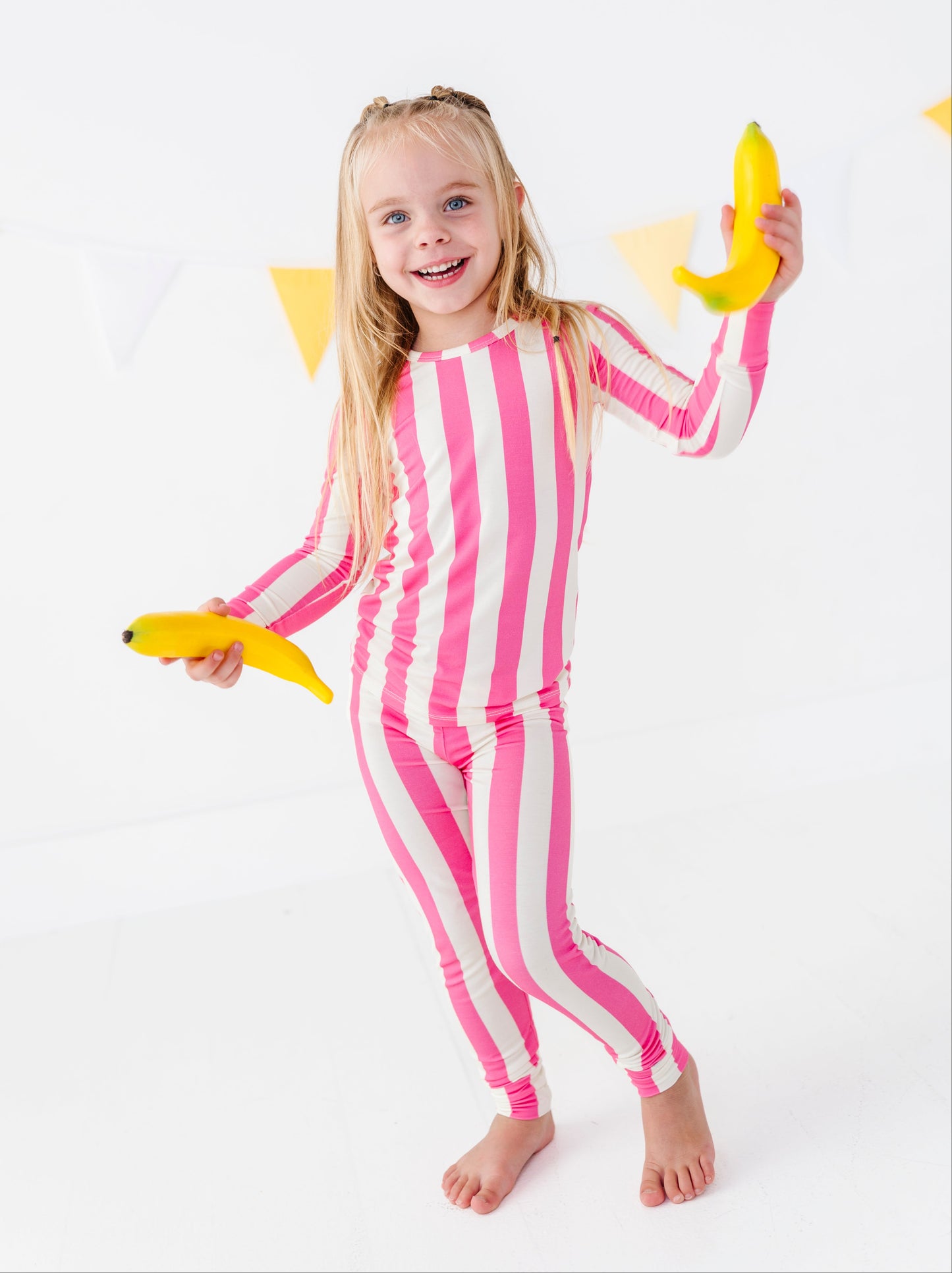 A young girl with blonde hair stands barefoot on a white studio background, wearing a pink and white vertically striped long-sleeve one-piece outfit and holding two yellow bananas.