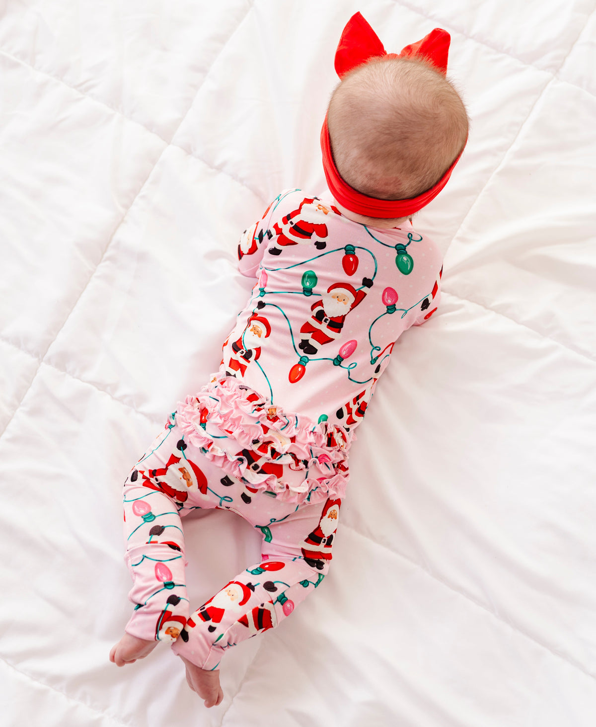 A detail shot of a baby girl wearing a pink ruffle bamboo convertible romper featuring  variety of Santas hanging from multicolor Christmas lights 