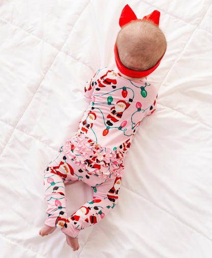 A detail shot of a baby girl wearing a pink ruffle bamboo convertible romper featuring  variety of Santas hanging from multicolor Christmas lights 
