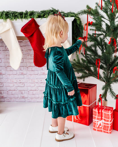 A young toddler girl in a dark green velvet tiered dress stands next to a decorated Christmas tree, touching red ornaments. The scene includes a white brick fireplace with red and white stockings, wrapped gifts, and a green garland.