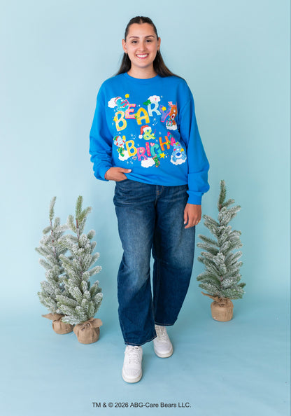 A young girl stands against a light blue studio backdrop wearing a bright blue crewneck sweatshirt with a colorful 'Beast Biker' graphic, dark blue jeans, and white sneakers. Two small flocked Christmas trees are placed on either side of her.