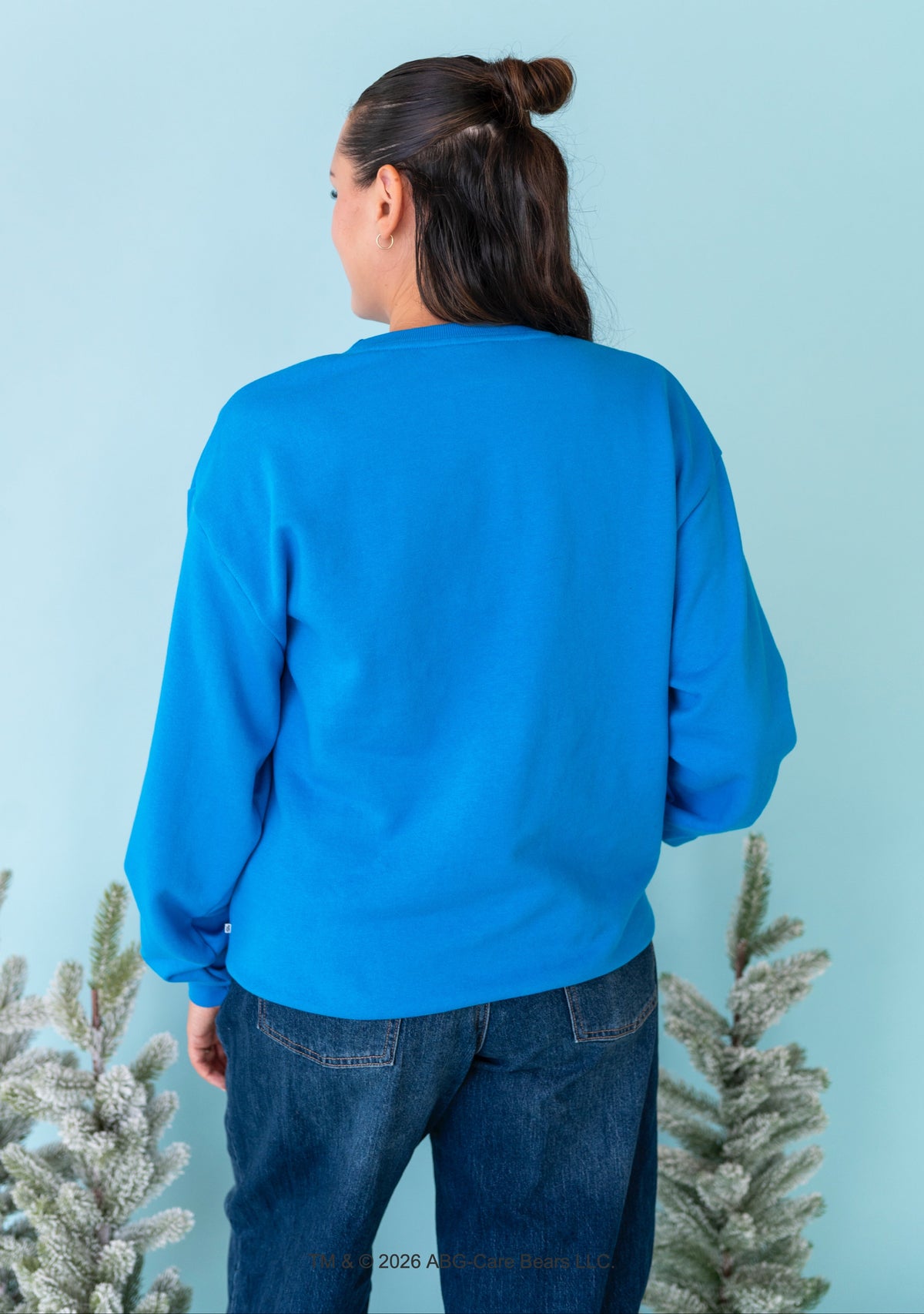 A woman viewed from behind wearing a bright blue long-sleeve sweatshirt and dark blue jeans, standing against a light blue background with frosted evergreen branches at the bottom corners.