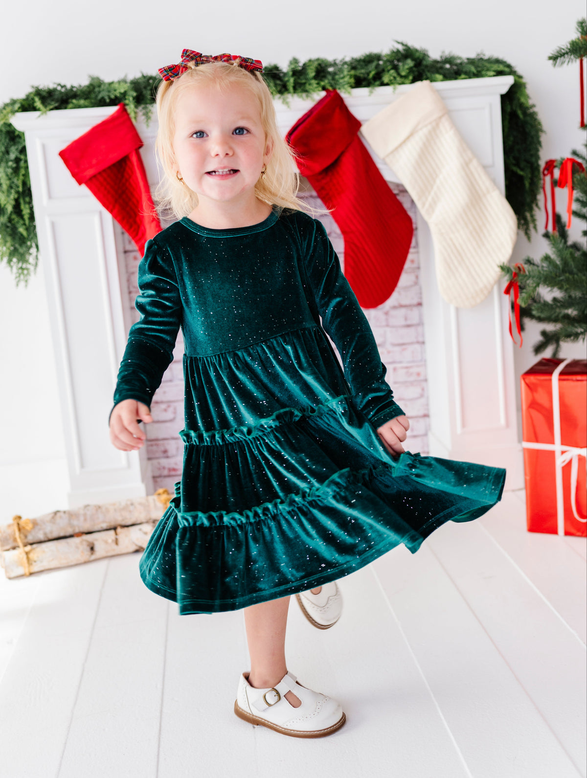 A young blonde girl wearing a sparkling dark green velvet long-sleeve dress with a ruffled hem, twirling in front of a decorated white fireplace with red and cream stockings.