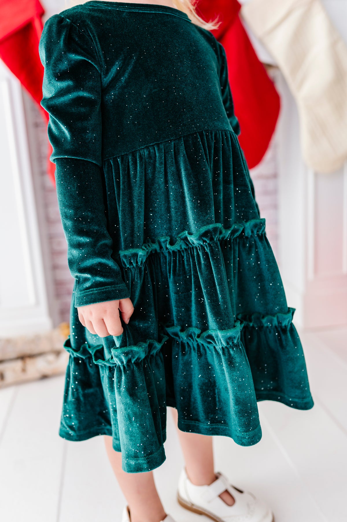 A toddler wearing a dark green velvet tiered dress with subtle silver glitter speckles, paired with white shoes, shown from the back in an indoor setting.