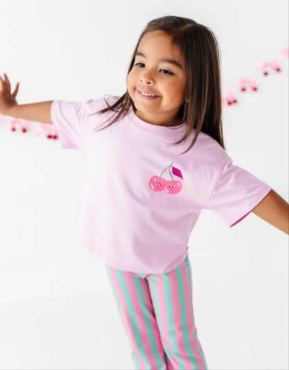 A young girl with long brown hair is smiling and posing with her arms outstretched, wearing a light pink short-sleeve t-shirt with a cherry graphic on the chest and pink and mint green striped pajama pants against a white background.