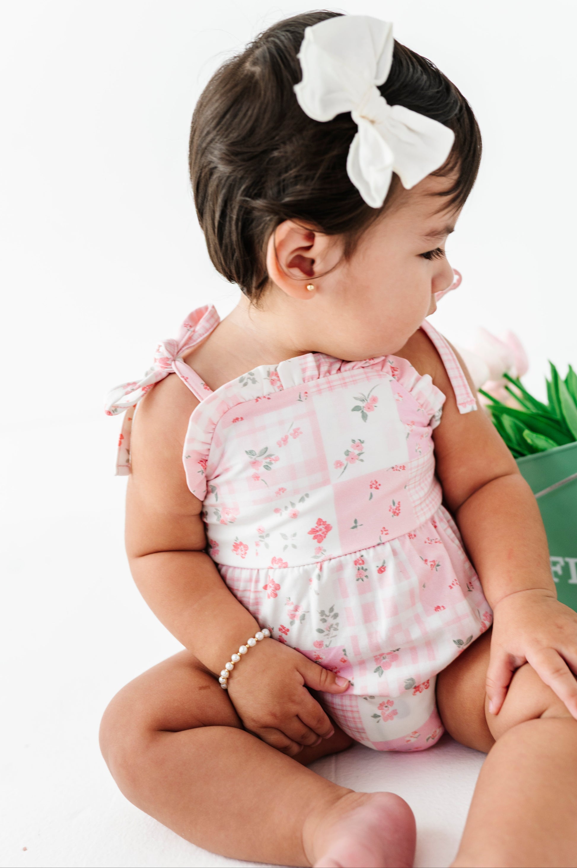 Baby in a pink floral bamboo bubble romper with a white bow sitting on a white surface.