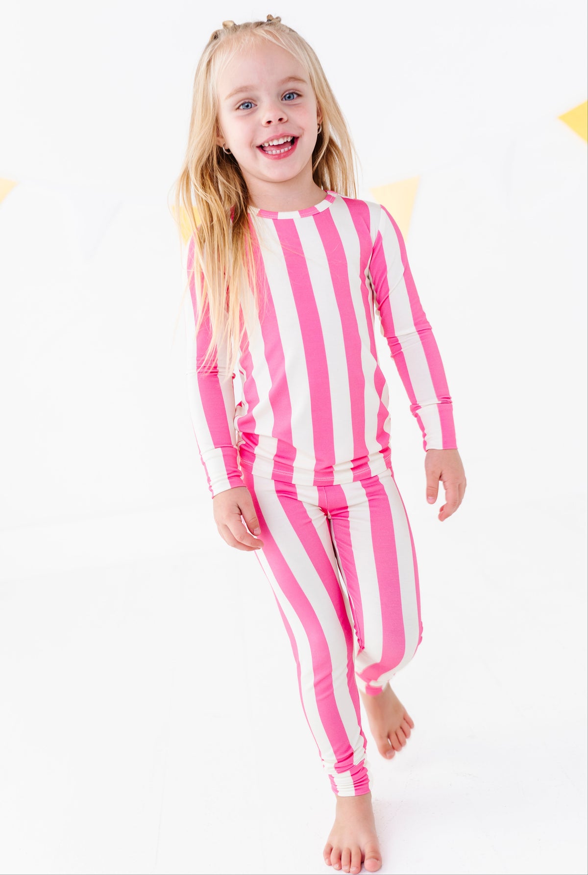 A young girl with blonde hair wearing a pink and white vertically striped long-sleeve rash guard and matching striped swim trunks, standing against a white background.