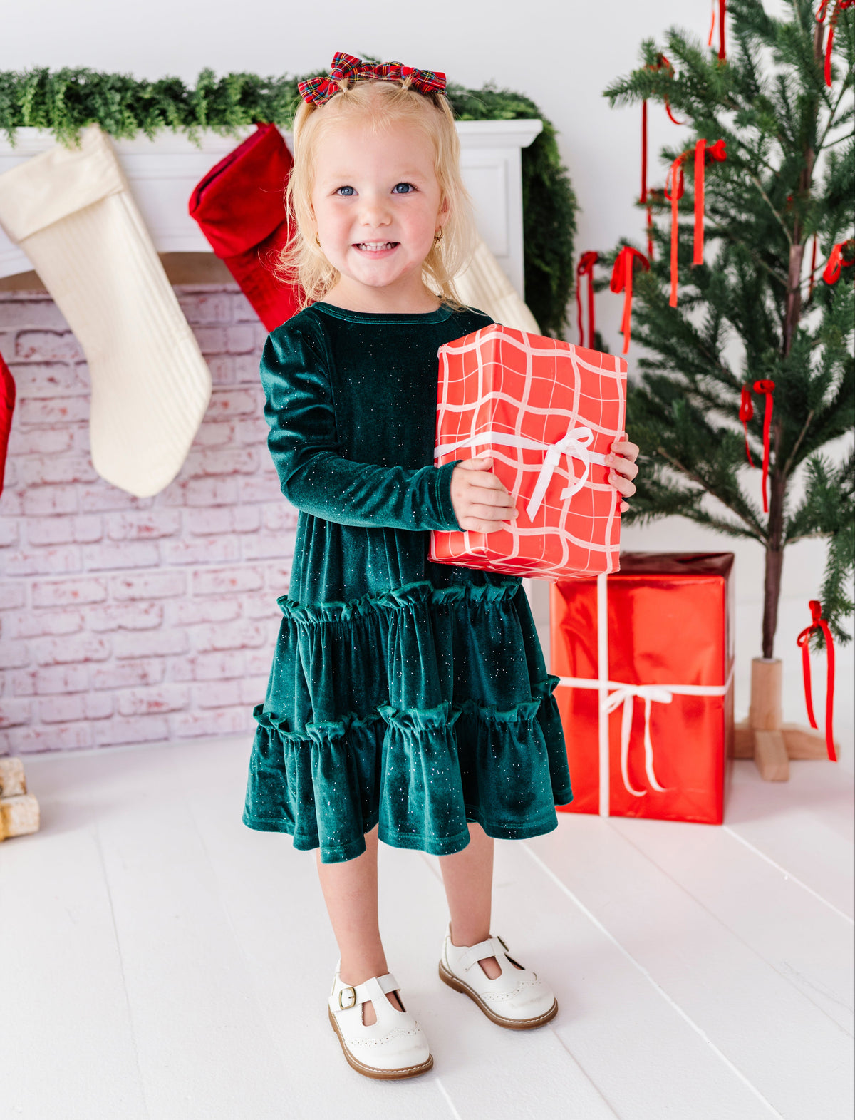 A young girl stands in a holiday-themed room wearing a dark green velvet tiered dress and holding a red gift box with white wrapping paper. A Christmas tree, stockings, and fireplace are visible in the background.