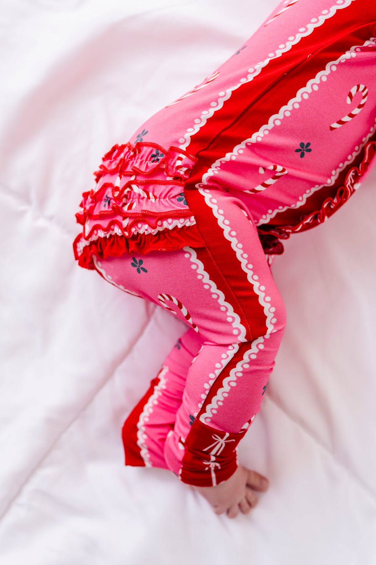 A side view of a baby girl wearing a red-and-pink bamboo Christmas convertible romper featuring ruffle detailing and foldover cuffs 