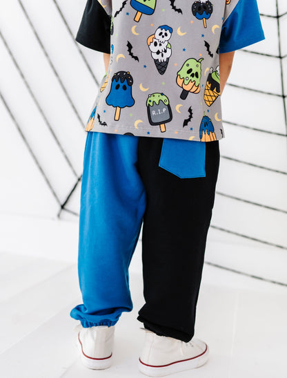 A child wearing a gray short-sleeve t-shirt with a colorful Halloween-themed print (ghost popsicles, skulls, bats) and split-color pants, one leg in bright blue and the other in black, paired with white sneakers.