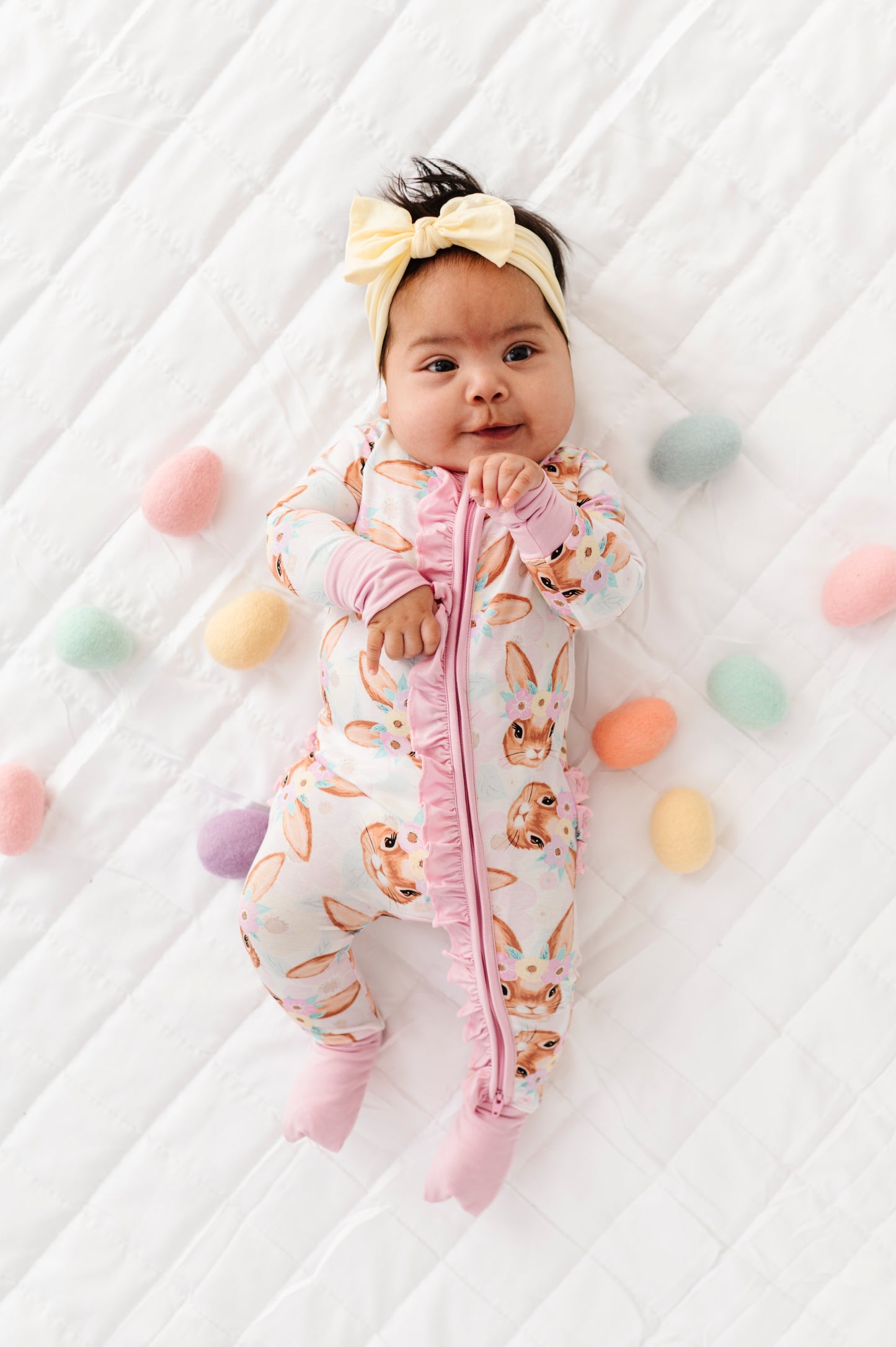 Baby in a bunny-themed onesie surrounded by colorful eggs on a white background