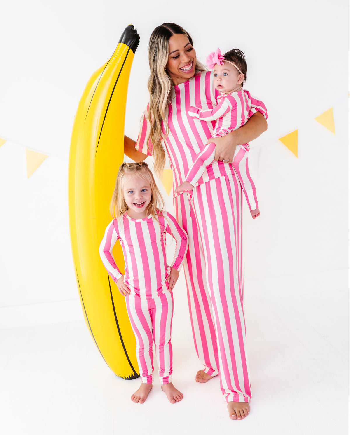 A woman and two young girls wearing matching pink and white vertically striped long-sleeve pajamas pose together in a studio setting. The woman holds a baby girl wearing a matching pink headband.