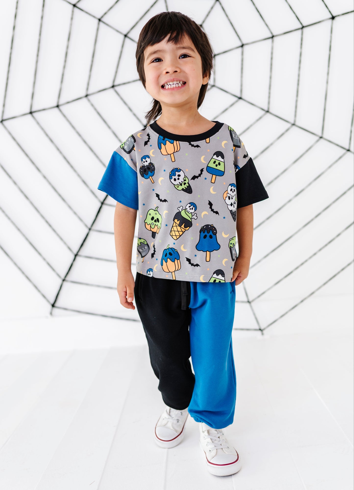 A young child stands in a studio wearing a short-sleeve t-shirt with a gray body covered in colorful Halloween-themed graphics (ghosts, pumpkins, bats) and contrasting blue and black sleeves, paired with black and blue pants and white sneakers. The background is white with a large black spiderweb graphic.