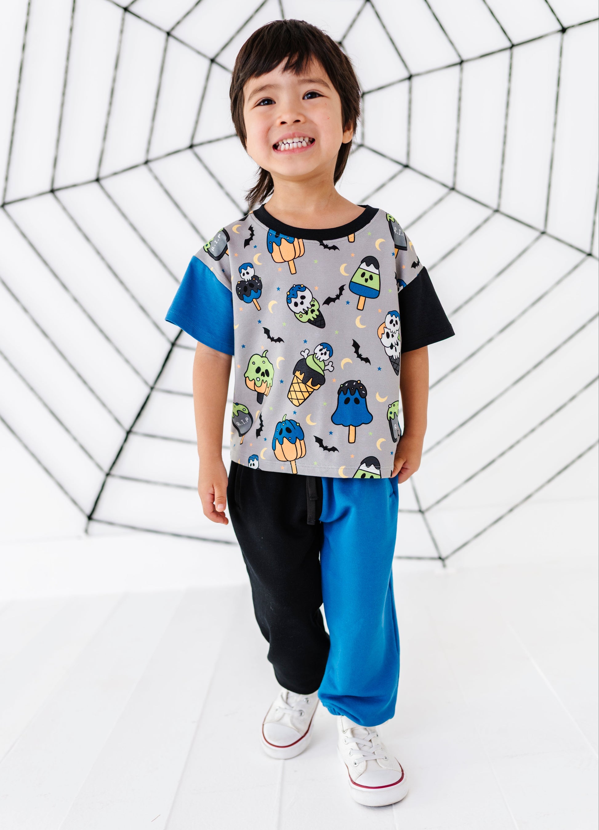 A young child stands in a studio wearing a short-sleeve t-shirt with a gray body covered in colorful Halloween-themed graphics (ghosts, pumpkins, bats) and contrasting blue and black sleeves, paired with black and blue pants and white sneakers. The background is white with a large black spiderweb graphic.