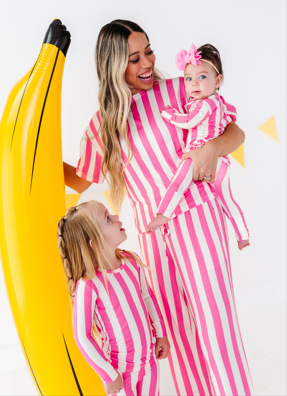 A woman and two children wearing matching pink and white striped one-piece outfits pose with a giant yellow inflatable banana prop against a white studio background.