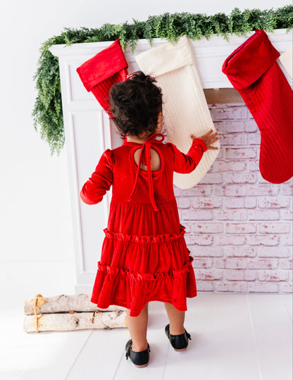 A toddler viewed from behind wearing a bright red long-sleeve ruffled dress, standing in front of a white fireplace mantel decorated with green garland, red and cream stockings, and a white brick pattern wall.