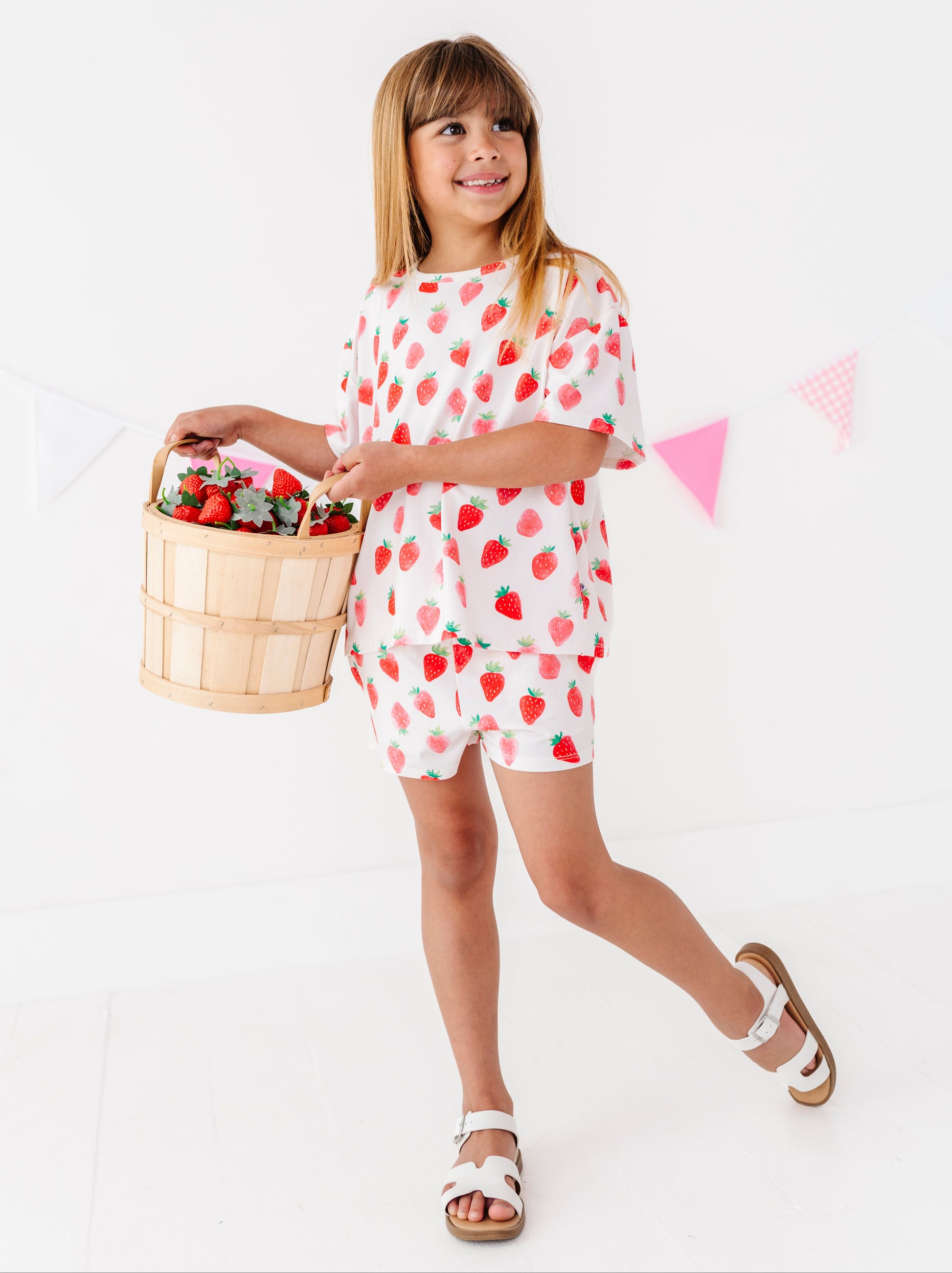 Young girl in a strawberry-patterned outfit holding a basket of flowers on a white background