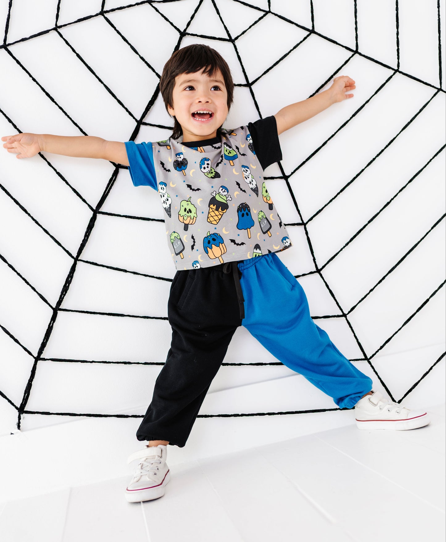 A young child wearing a gray t-shirt with a colorful owl pattern, a black and blue long-sleeve top, and black and blue pants, posing with arms outstretched against a white wall with a black spiderweb design.