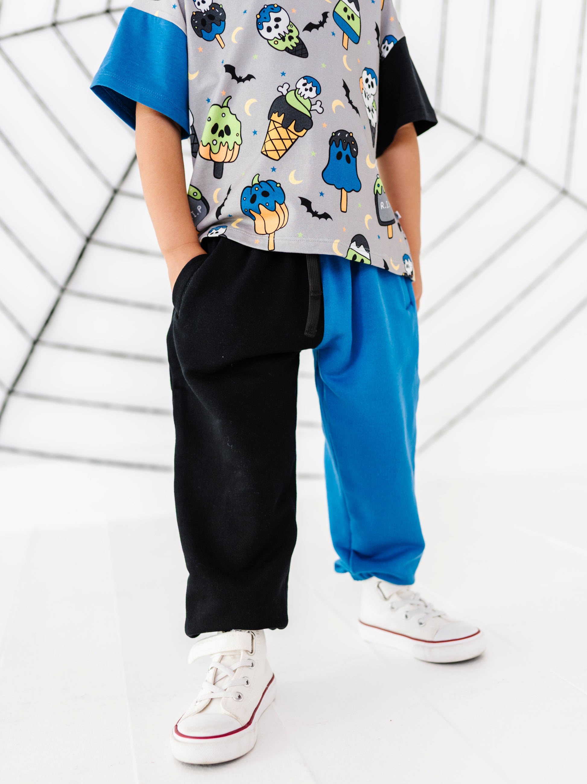 A child wearing a short-sleeve t-shirt with a colorful Halloween ice cream pattern, featuring split black and blue sweatpants, and white sneakers, standing against a white background with a spiderweb graphic.