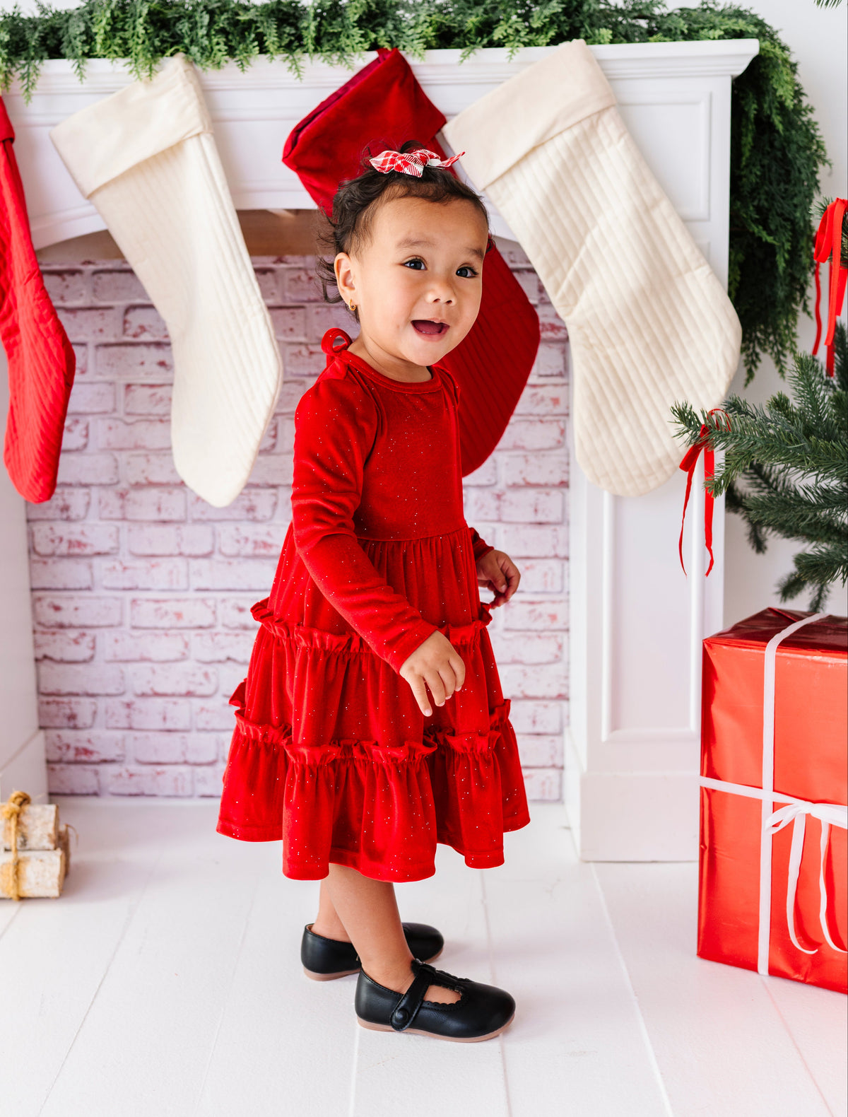 A toddler girl wearing a festive red long-sleeve dress and black shoes stands in front of a white fireplace mantel decorated with red and cream stockings, green garland, and a wrapped red gift box.