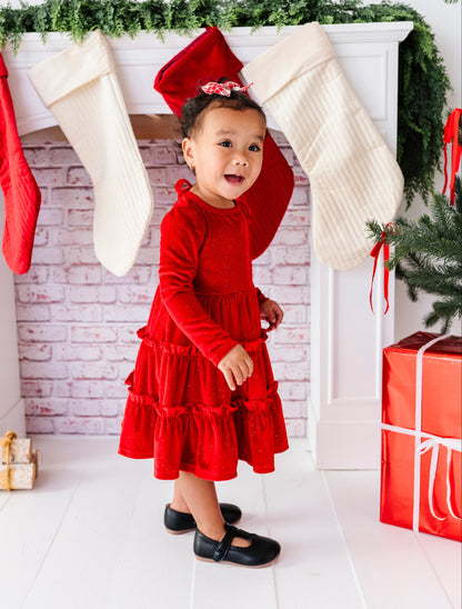 A toddler girl wearing a festive red long-sleeve dress and black shoes stands in front of a white fireplace mantel decorated with red and cream stockings, green garland, and a wrapped red gift box.