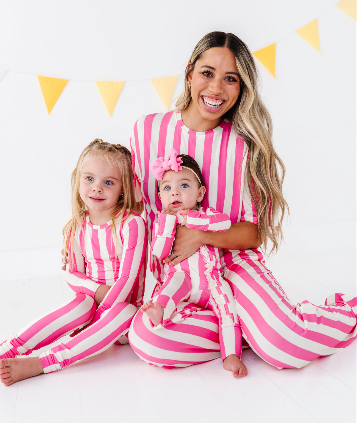 A woman and two young girls wearing matching pink and white vertically striped long-sleeve pajamas sit together on a white floor against a white wall with yellow bunting.