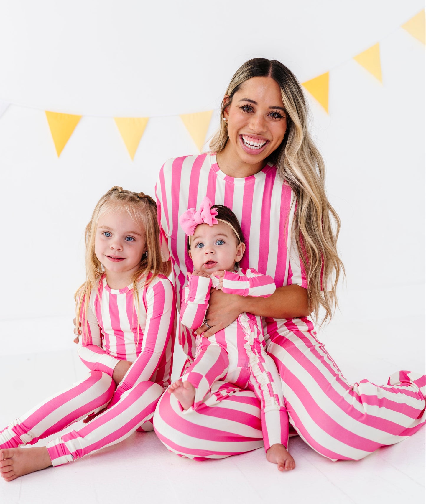A woman and two young girls wearing matching pink and white vertically striped long-sleeve pajamas sit together on a white floor against a white wall with yellow bunting.
