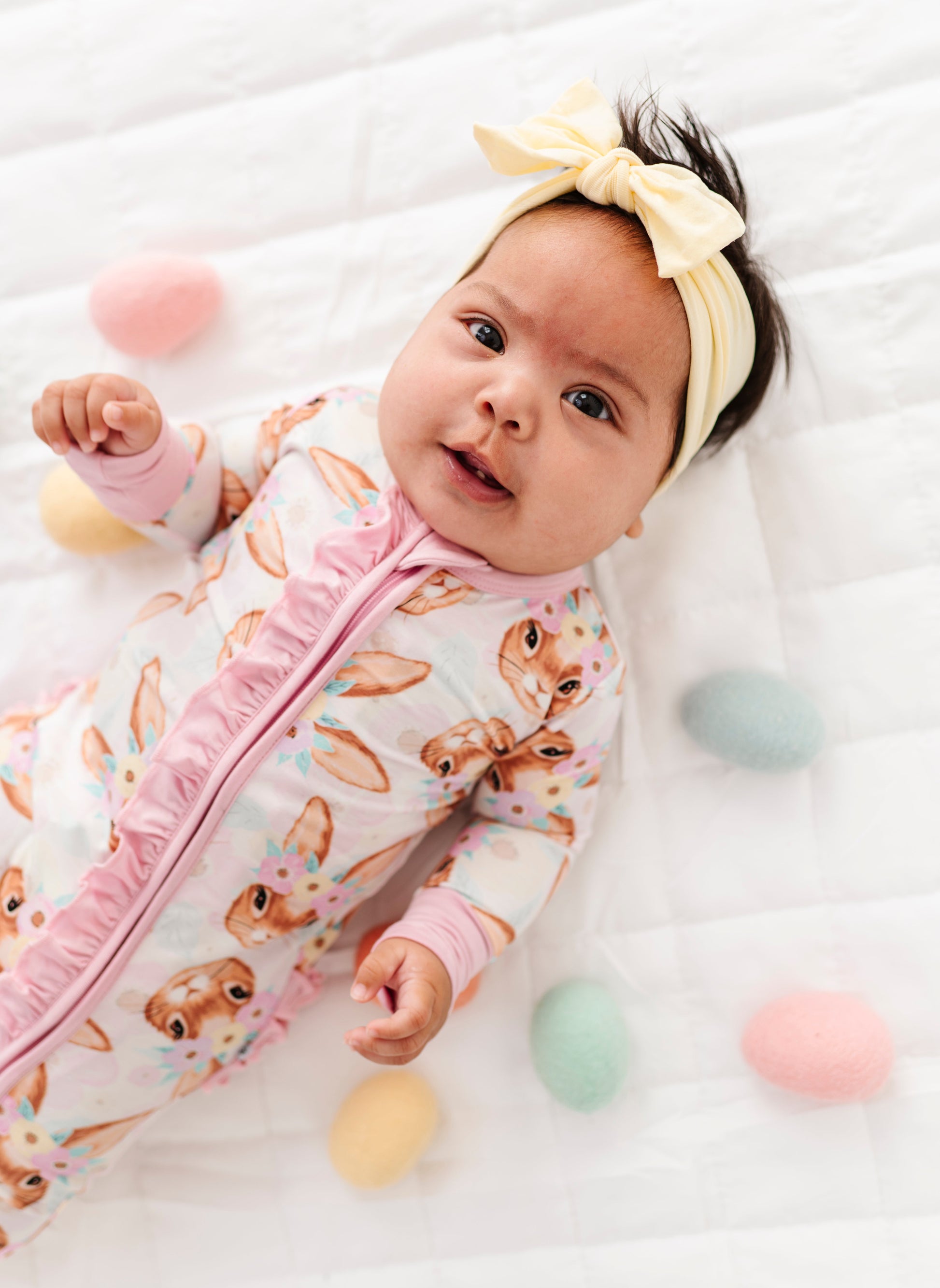 Baby lying on a white blanket with colorful eggs and a yellow headband.