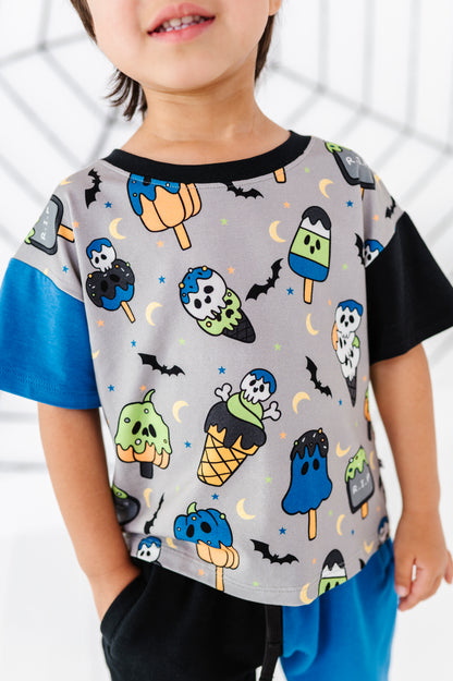 A child wearing a short-sleeve t-shirt with a gray body covered in a Halloween ice cream cone and popsicle print, with solid black sleeves and collar, and bright blue side panels.