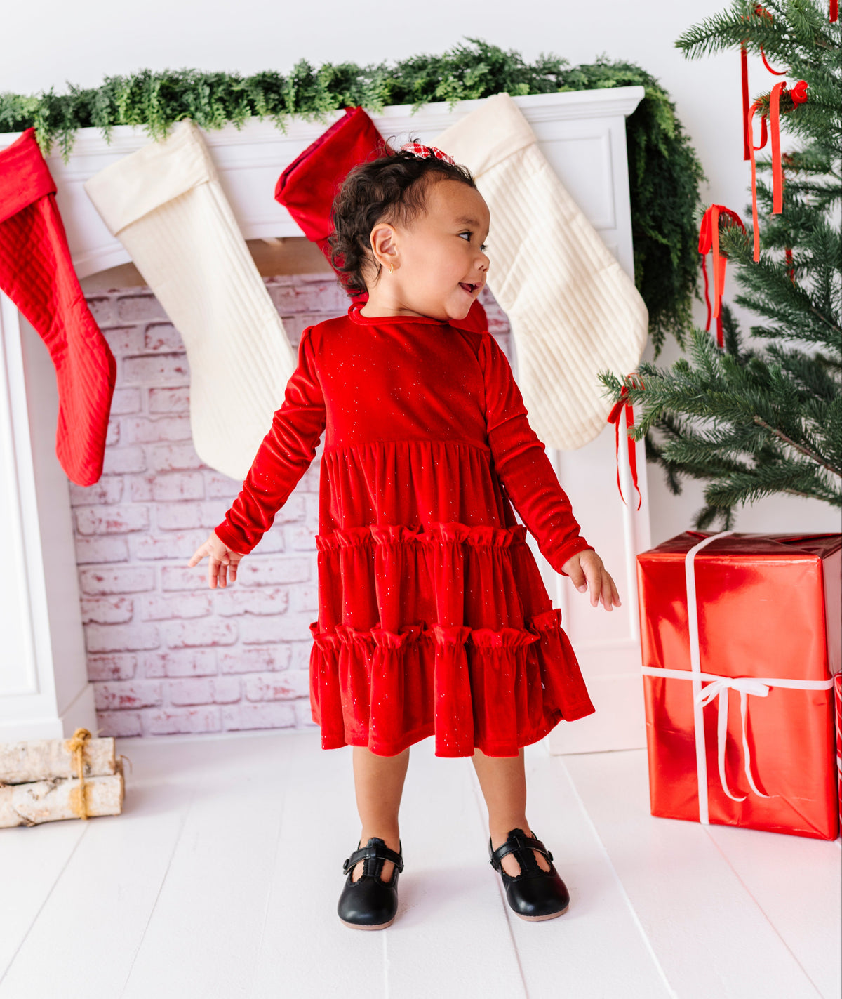 A toddler girl wearing a festive red velvet long-sleeve tiered dress stands in front of a decorated Christmas fireplace with stockings, a tree, and wrapped presents.