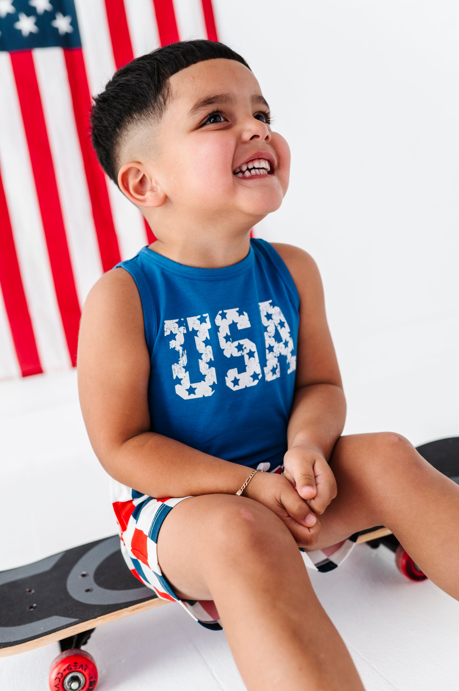 Child wearing a bamboo-cotton 'USA' tank top sitting on a skateboard with an American flag in the background